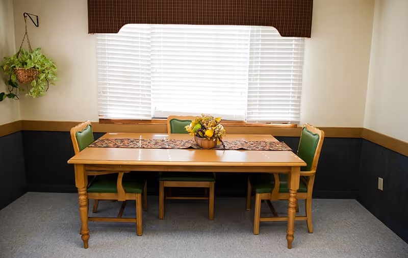 A small dining room with a wooden table, four green-cushioned chairs, a fruit centerpiece and a window with closed blinds.