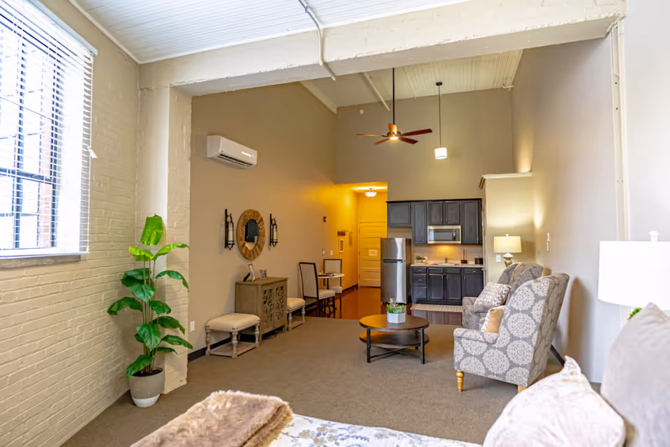 A spacious, well-lit living area with high ceilings featuring a ceiling fan and pendant light. The room includes a small kitchen with dark cabinets, a stainless steel refrigerator, microwave, and sink. There are two patterned armchairs with cushions, a round wooden coffee table with a small plant, and a console table with a round mirror and decorative items. A large window with blinds and a potted plant are visible on the left side. The walls are painted beige, and the floor is carpeted.