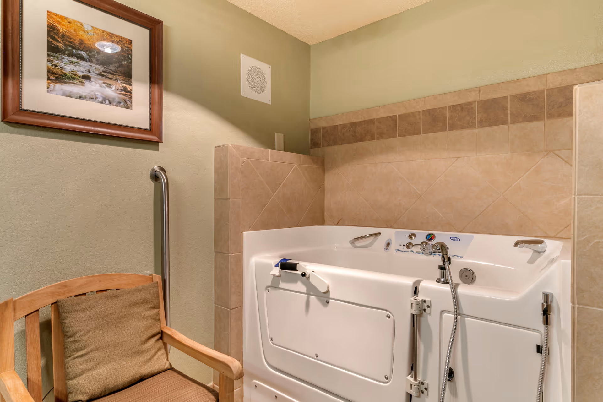A bathroom featuring a walk-in bathtub with safety handles and controls, beige tiled walls, a wooden chair with a cushion, and a framed picture of a forest stream on the wall.