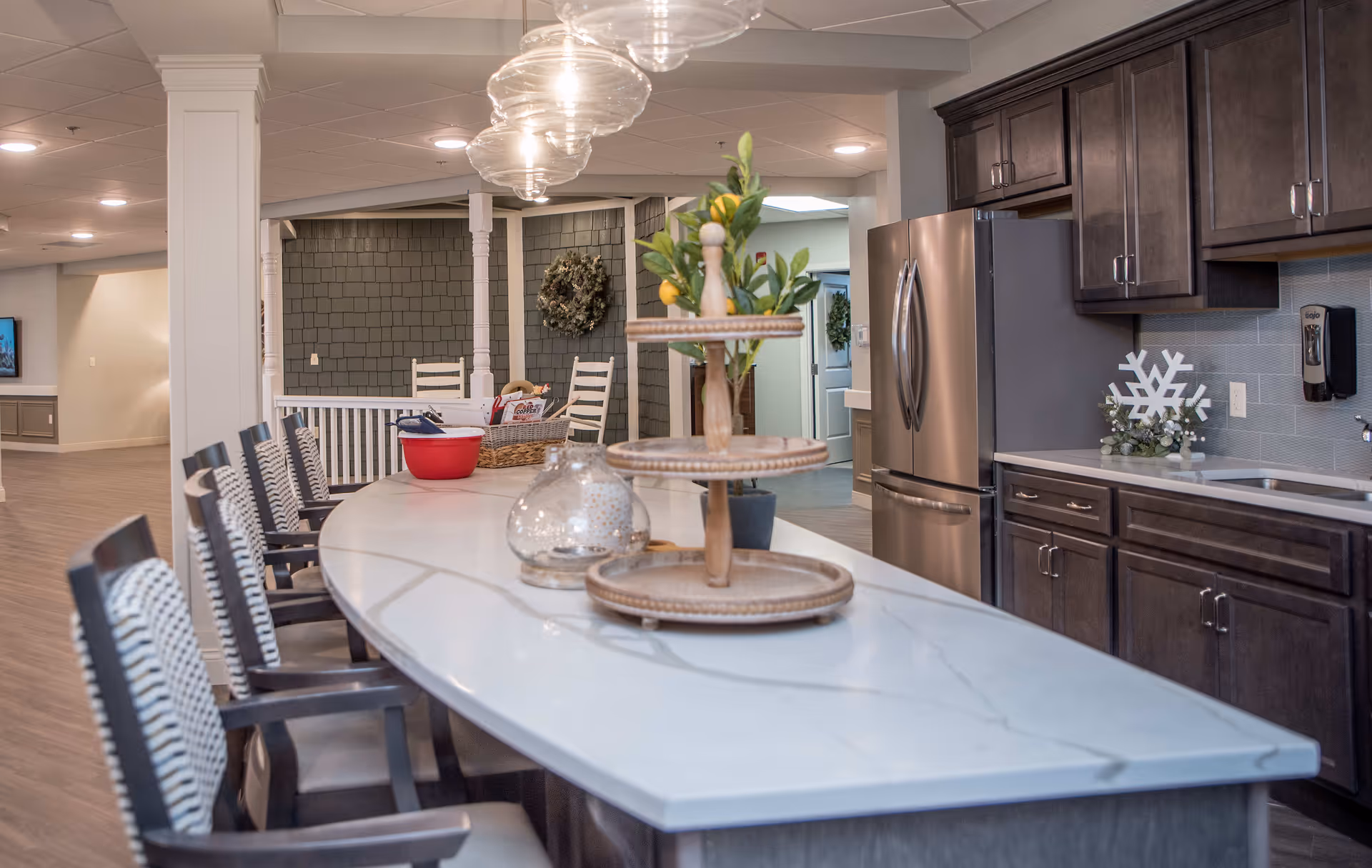 Modern communal kitchen with a long marble island, bar stools, pendant lights, stainless refrigerator, and decorative centerpiece.