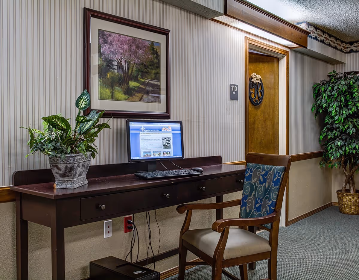 A hallway area in a senior living facility featuring a dark wooden desk with a computer monitor, keyboard, and mouse. A potted plant is placed on the desk. There is a wooden chair with a patterned blue cushion in front of the desk. The wall has striped wallpaper with a framed painting of a tree with pink blossoms. A door labeled 110 with a decorative wreath is visible, along with a large potted artificial tree in the corner.