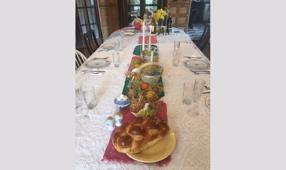 A long dining table set with a white lace tablecloth and a colorful table runner. The table is arranged with plates, glasses, cutlery, and decorative items including baskets, flowers, candles, and a loaf of braided bread on a plate at the near end. The room has wooden furniture and a window in the background.