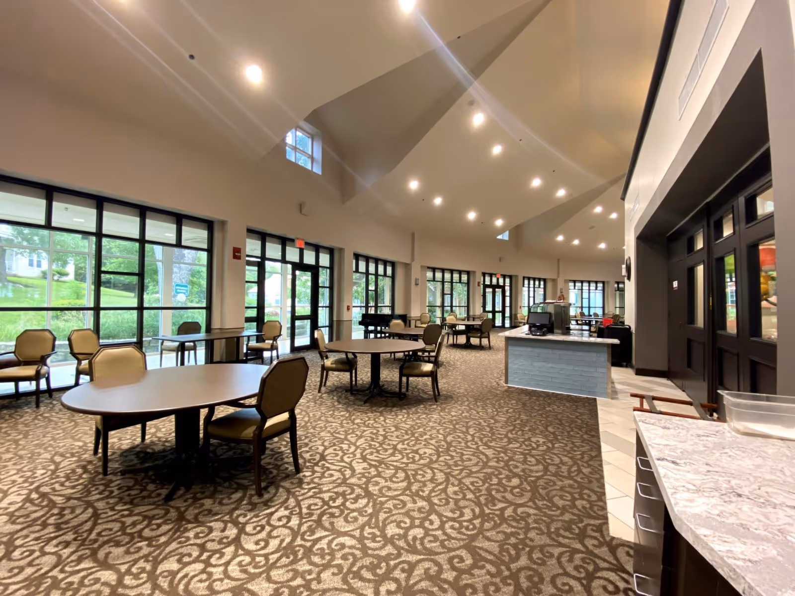 Spacious dining area with multiple round tables and chairs arranged on a patterned carpet. Large windows along the walls allow natural light to fill the room, and there is a counter area with a marble countertop on the right side. The ceiling is high with recessed lighting.