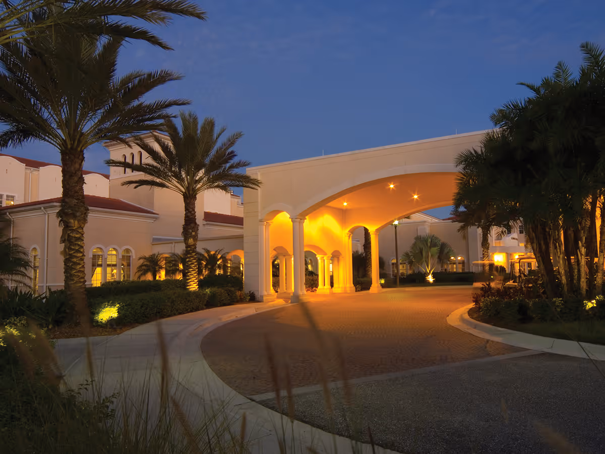 Evening view of the entrance to The Glenridge on Palmer Ranch, featuring a large covered driveway with arches and columns, surrounded by palm trees and well-lit landscaping.