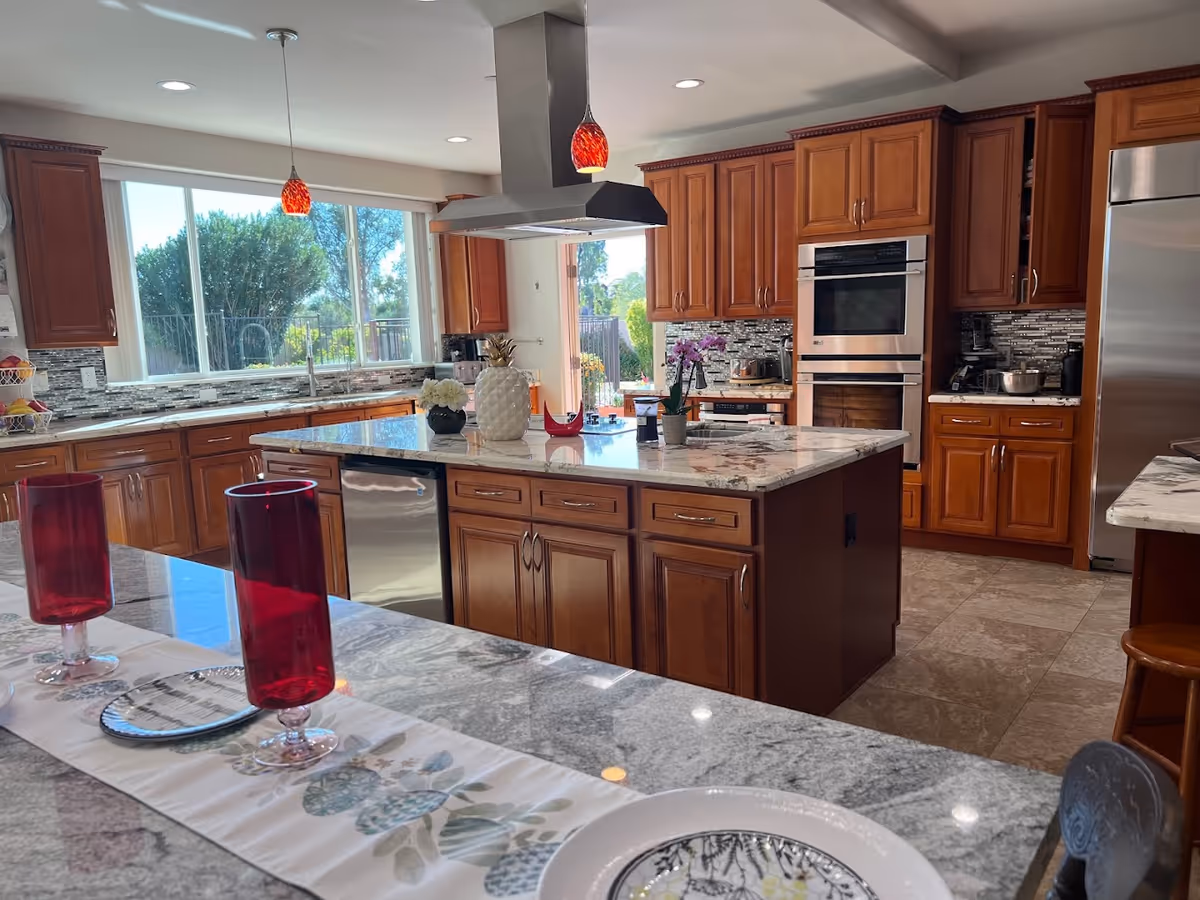 Spacious kitchen with a large central island, wooden cabinets, marble countertops, stainless steel appliances and red glassware on the foreground counter.