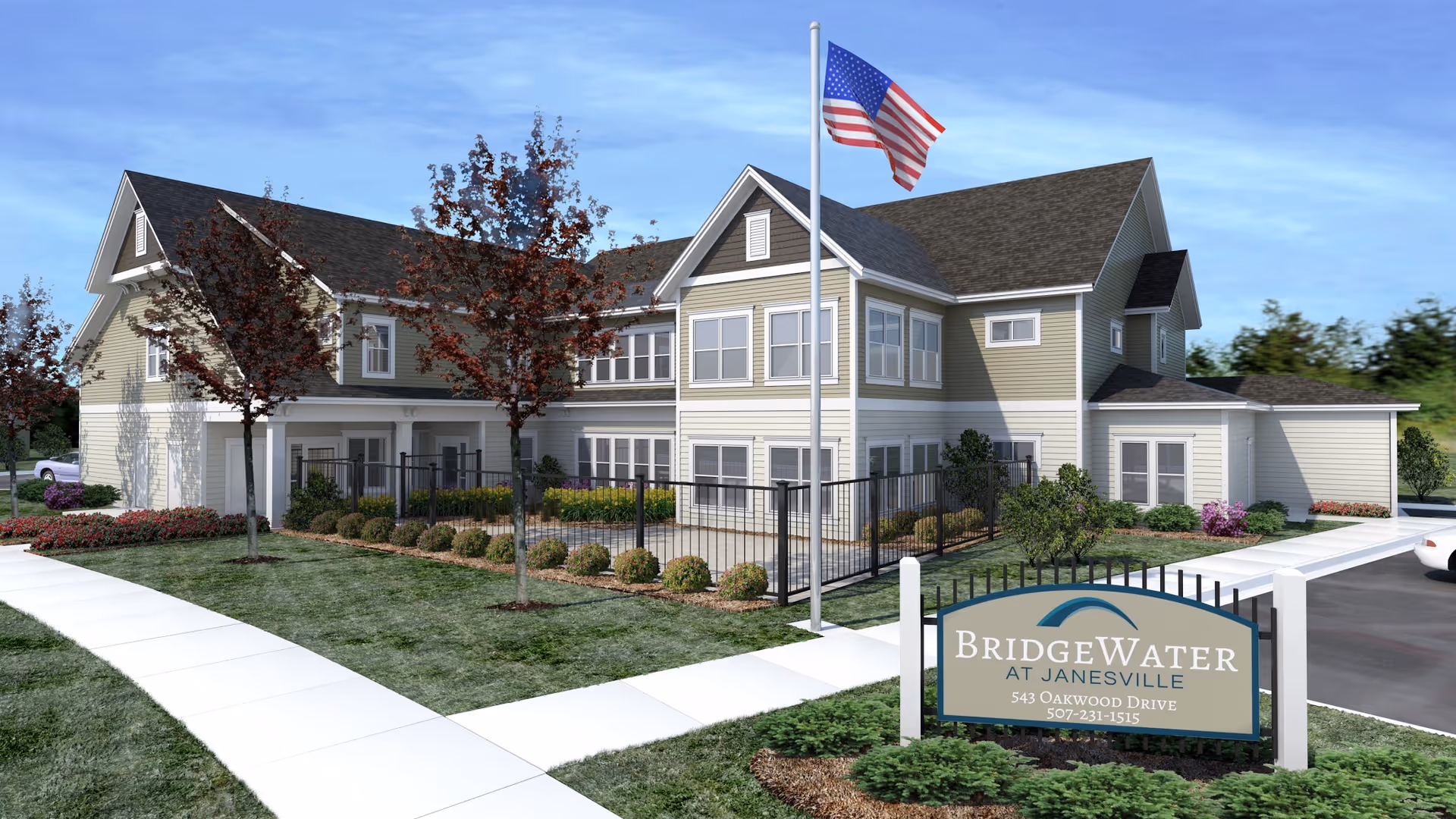 Exterior view of BridgeWater at Janesville, a two-story senior living facility with beige siding and white trim. The building is surrounded by landscaped greenery, small trees, and a sidewalk. An American flag flies on a flagpole near the entrance, and a sign in front displays the facility's name and contact information.