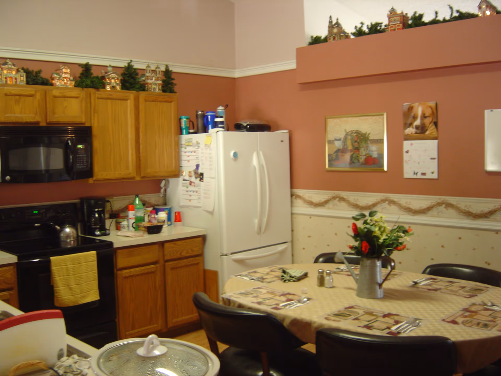 A combined kitchen and dining area with oak cabinets, a white refrigerator and stove, and a round table set with placemats and a flower centerpiece.
