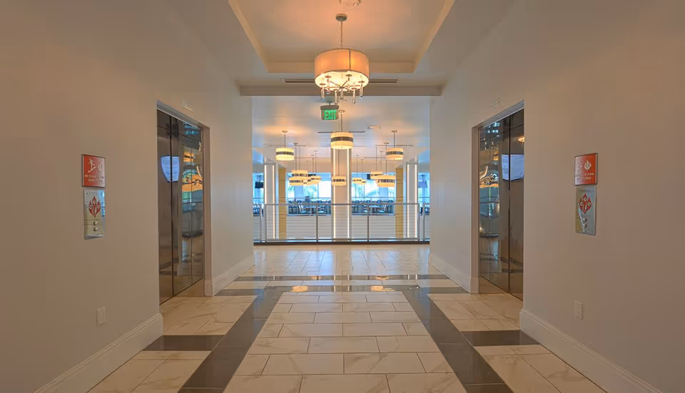A hallway with two elevators on either side, leading to a bright dining area with multiple hanging light fixtures and tables visible in the background. The floor is tiled with a pattern of white and dark tiles, and there are fire safety signs next to each elevator.