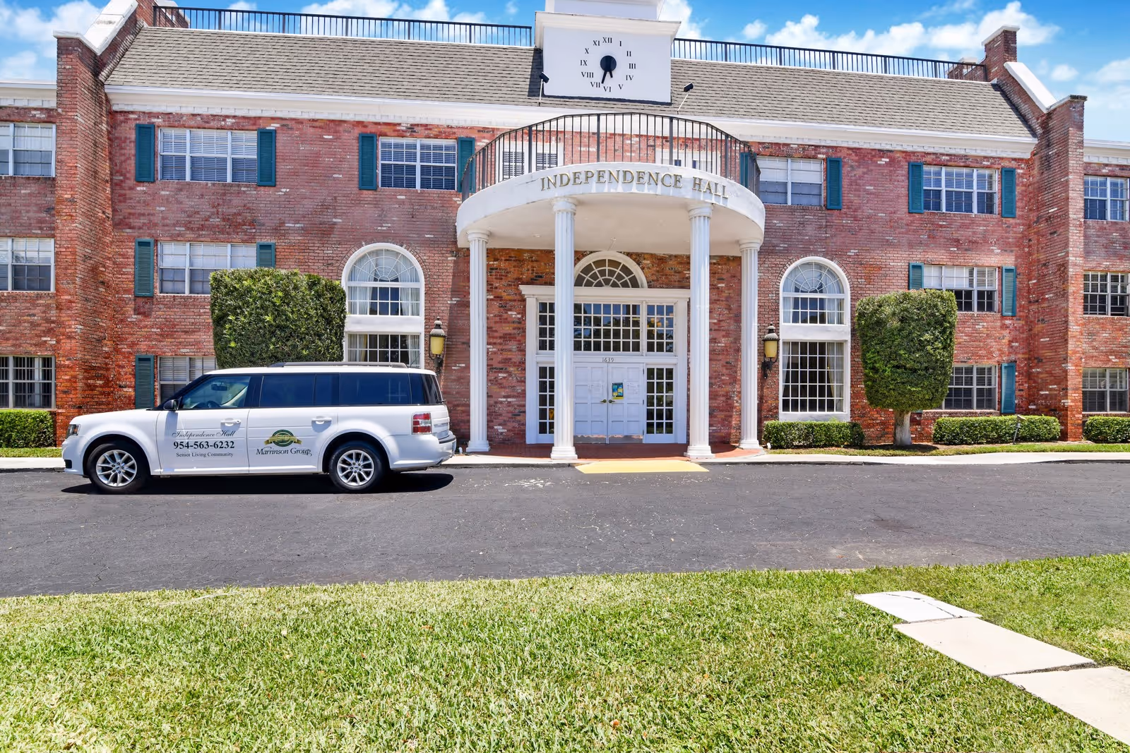 Front exterior view of a red brick building named Independence Hall with white columns at the entrance and a clock above the entrance. A white vehicle with signage for Morrison Group and a phone number is parked in front of the building. The scene includes a paved driveway and green grass in the foreground under a blue sky with some clouds.