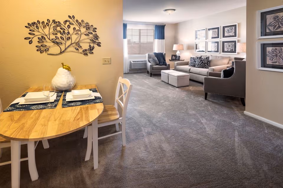 A cozy living space featuring a small wooden dining table set for two with white chairs and a decorative pear centerpiece. The room extends to a living area with a beige sofa, two armchairs, ottomans, side tables with lamps, and framed floral artwork on the walls. The space is carpeted and well-lit by natural light from a window with blue and white curtains.