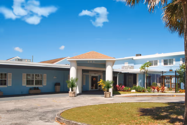 Exterior front view of Haines Manor Assisted Living facility showing a light blue building with white columns at the entrance, a small garden area with plants, and a clear blue sky with some clouds.