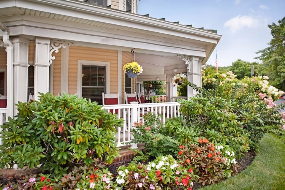 Front porch of a building with white railings and decorative trim, featuring several rocking chairs with red cushions. The porch is surrounded by lush green bushes and colorful flowers, with a hanging basket of yellow flowers. The sky is partly cloudy and an American flag is visible in the background.