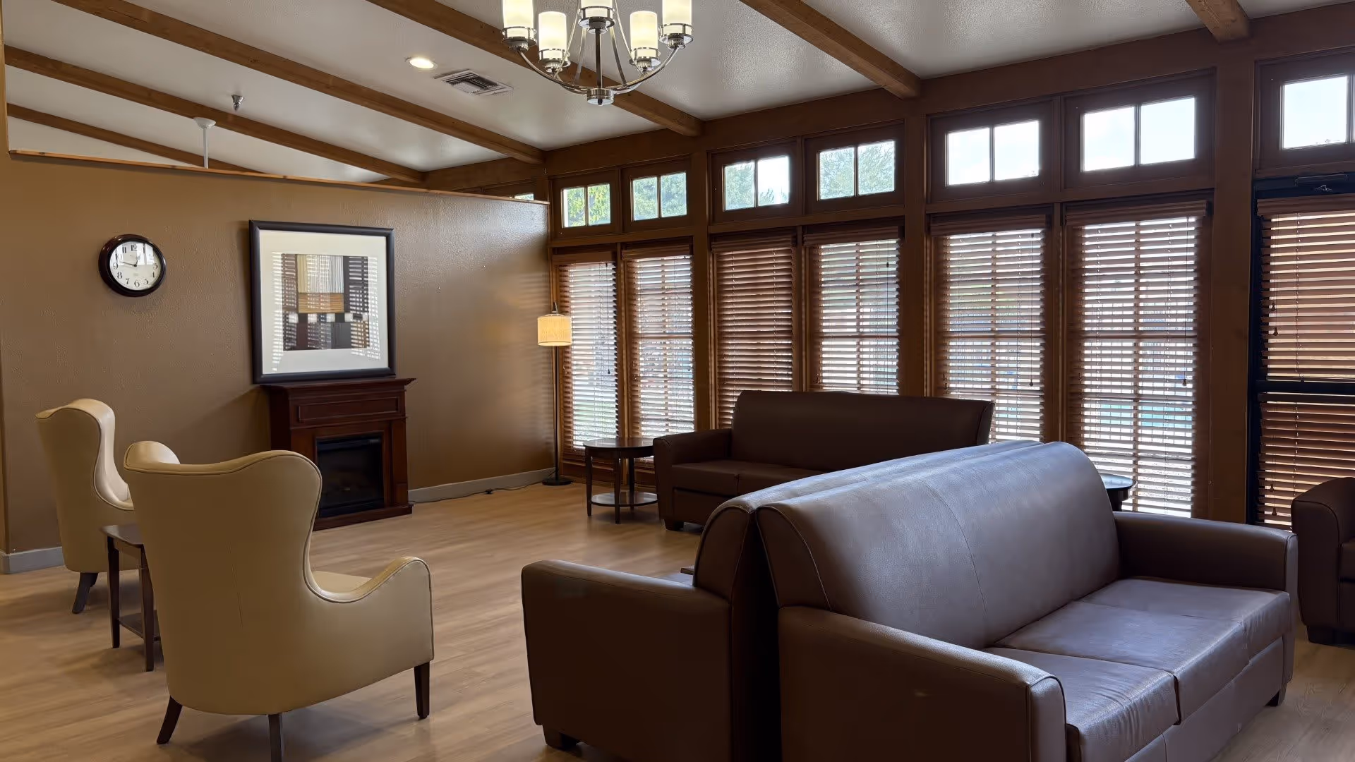 A cozy living room area with brown leather sofas and beige armchairs arranged around a small wooden table. The room features large windows with wooden blinds, a wooden floor, a wall clock, a framed picture above a fireplace, and a ceiling light fixture with multiple bulbs.