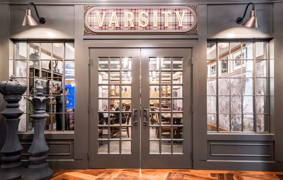 Entrance to a room labeled 'VARSITY' with double glass-paneled doors and windows on either side. Inside, people are seated at tables in a well-lit room with decorative wall art and wooden furniture. Large chess pieces are positioned to the left of the entrance.