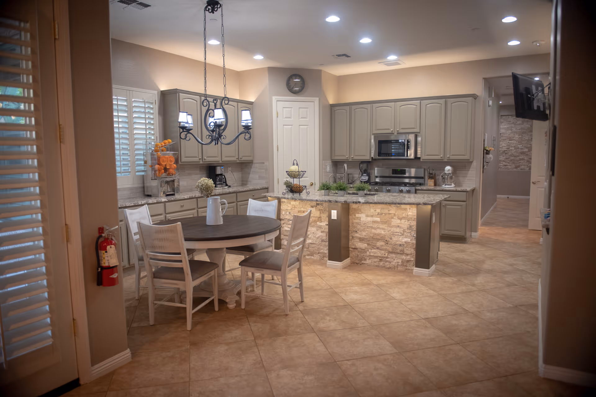 A spacious kitchen and dining area with beige tiled floors and light gray cabinets. The kitchen features a central island with a stone facade, stainless steel appliances including a microwave and stove, and a round wooden dining table with four white chairs. A decorative chandelier hangs above the table, and there are various kitchen items and plants on the counters. The room is well-lit with recessed ceiling lights and natural light coming through white plantation shutters on the window.