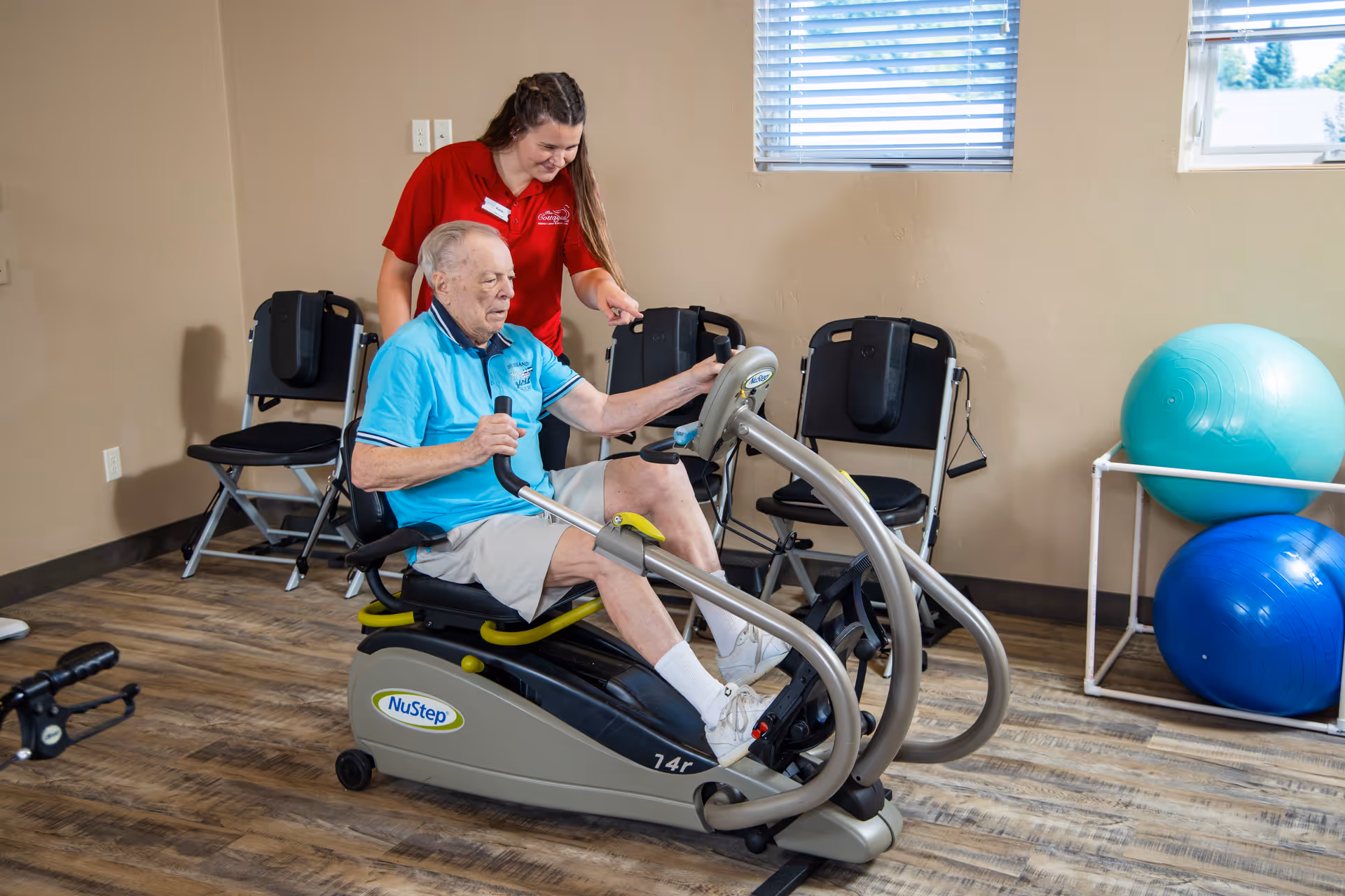 An elderly man using a NuStep exercise machine in a senior living facility's fitness room, assisted by a female staff member wearing a red shirt with the facility's logo. The room has wooden flooring, chairs against the wall, and exercise balls near a window with blinds.