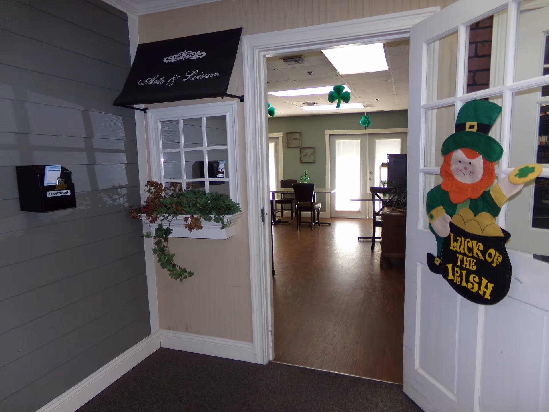View through a doorway into a communal activity room decorated for St. Patrick's Day with shamrocks and a 'Luck of the Irish' leprechaun on the door.
