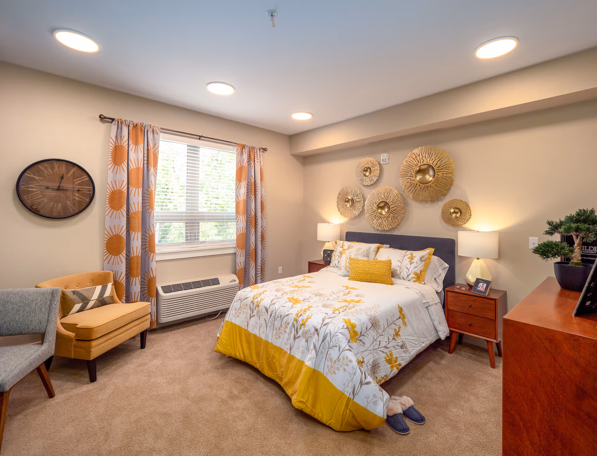 A cozy bedroom in a senior living community featuring a bed with floral and yellow bedding, two wooden nightstands with lamps, decorative wall art above the bed, a window with patterned curtains, a yellow armchair with a pillow, a gray chair, and a wooden dresser with a small plant and framed photo.