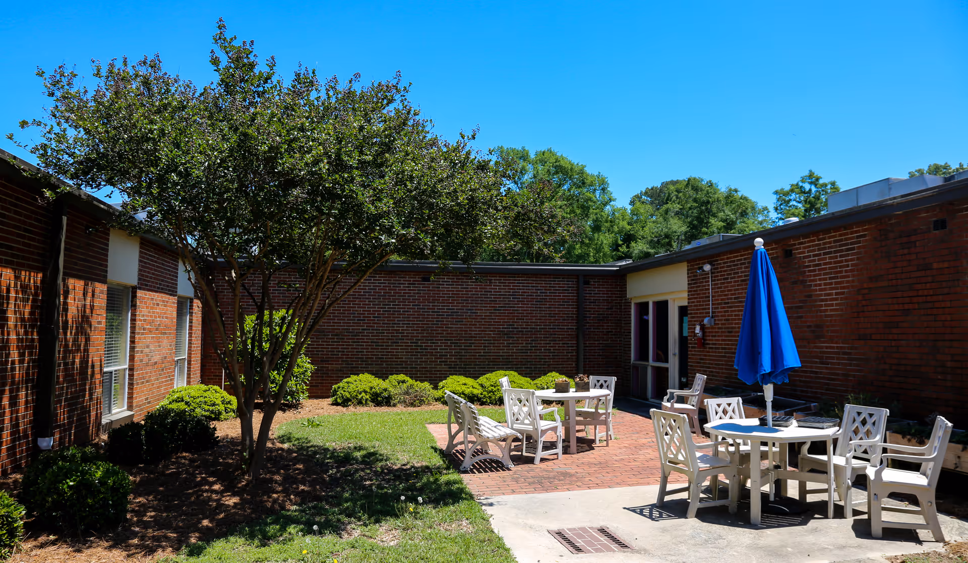 Sunny brick courtyard with patio tables and chairs, a blue umbrella, and landscaping.
