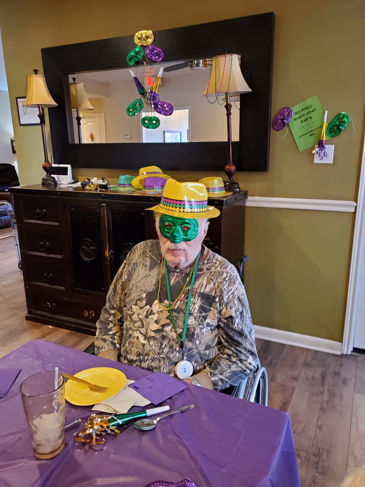 An elderly man wearing a green mask, yellow hat, and colorful beads sits in a wheelchair at a table covered with a purple tablecloth. The table has a yellow plate, utensils, a glass, and party decorations. Behind him is a dark wooden cabinet with lamps and more yellow hats on top. The wall has a large mirror and Mardi Gras decorations, including masks and a green sign that reads 'The Pines Mardi Gras Party'.