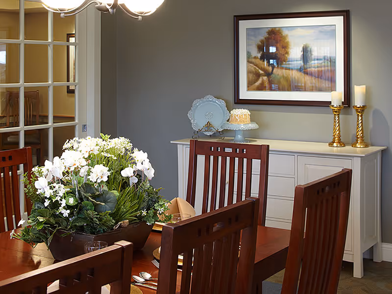 A dining room with a wooden table and matching chairs. The table is decorated with a large floral centerpiece. In the background, there is a white sideboard with decorative items including a cake on a stand, a plate on a stand, and two gold candlesticks with white candles. A framed landscape painting hangs on the gray wall above the sideboard. A chandelier with multiple lights hangs above the table.