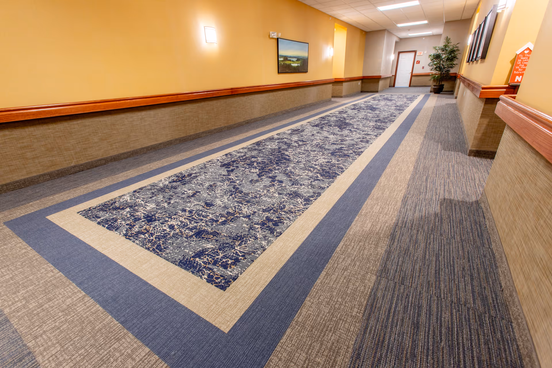 A long, well-lit hallway with beige and yellow walls, a patterned blue and beige carpet runner in the center, wooden chair rails, wall-mounted lights, framed artwork, and a potted plant near the end of the corridor.