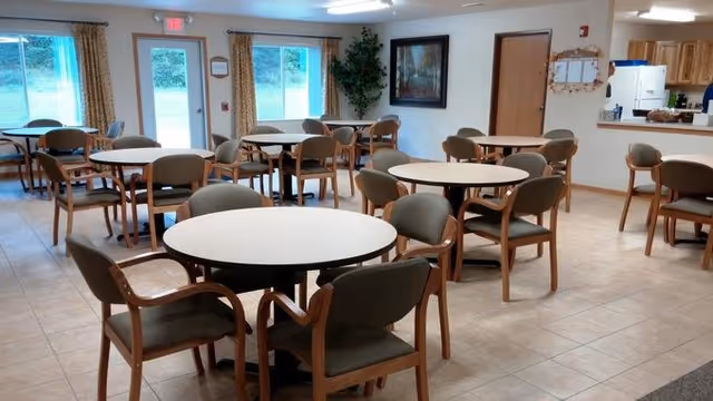Communal dining room with multiple round tables and wooden chairs, tiled floor, windows, and a serving counter in the background.