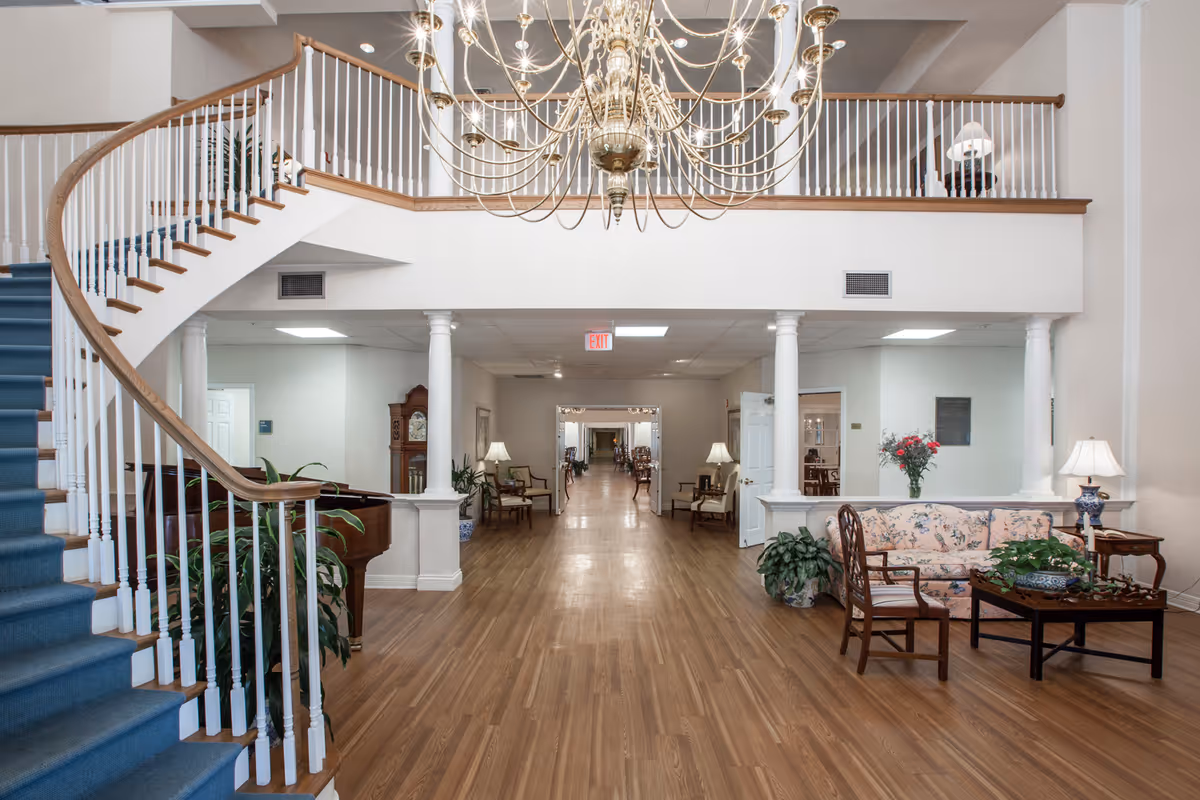 Spacious interior of a senior living facility featuring a curved staircase with blue carpet, wooden handrails, and white balusters. The room has wooden flooring, white walls, and a large brass chandelier hanging from the ceiling. There is a floral-patterned sofa, wooden chairs, a coffee table with plants, and a grand piano near the staircase. The space is decorated with lamps, plants, and a grandfather clock, with a long hallway visible in the background.