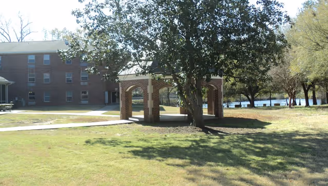 Grassy lawn with a brick arched pavilion beneath a large tree in front of a multi-story brick building and a pond.