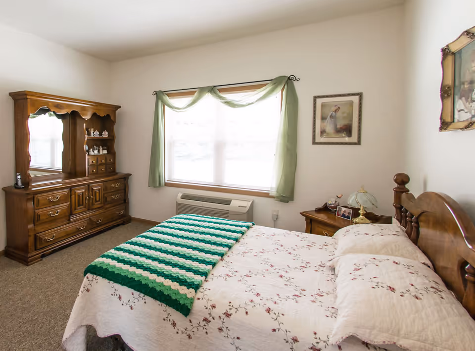 Furnished bedroom with a wooden dresser, queen bed, floral quilt, and green window curtains.