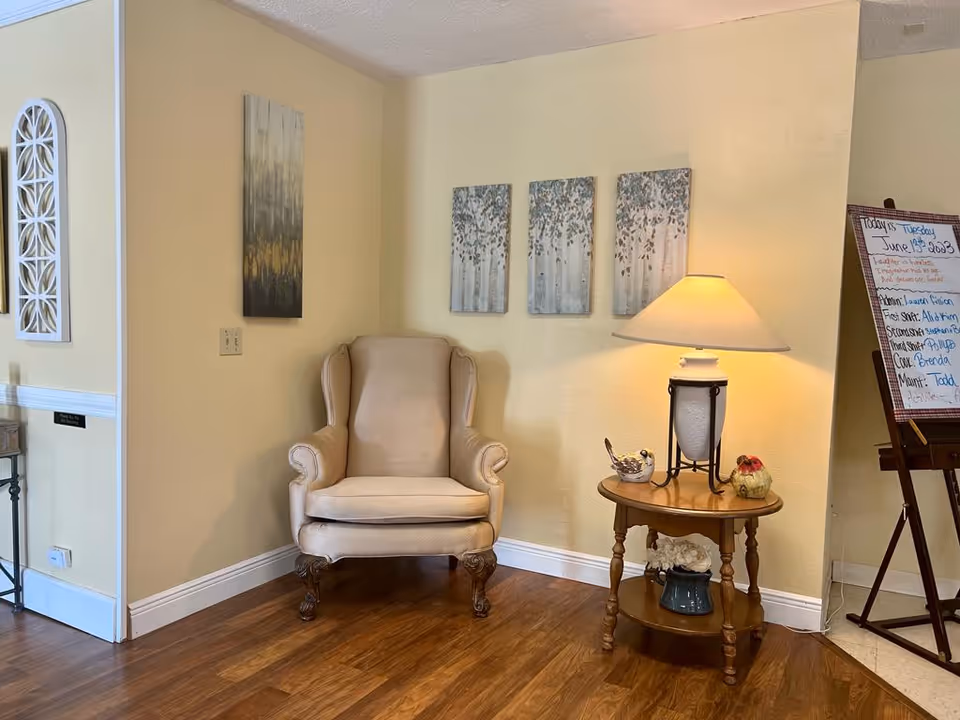 A cozy corner in a living area with a beige upholstered armchair, a wooden side table with a lamp and decorative items, three nature-themed paintings on the wall, and a whiteboard with handwritten notes on an easel.