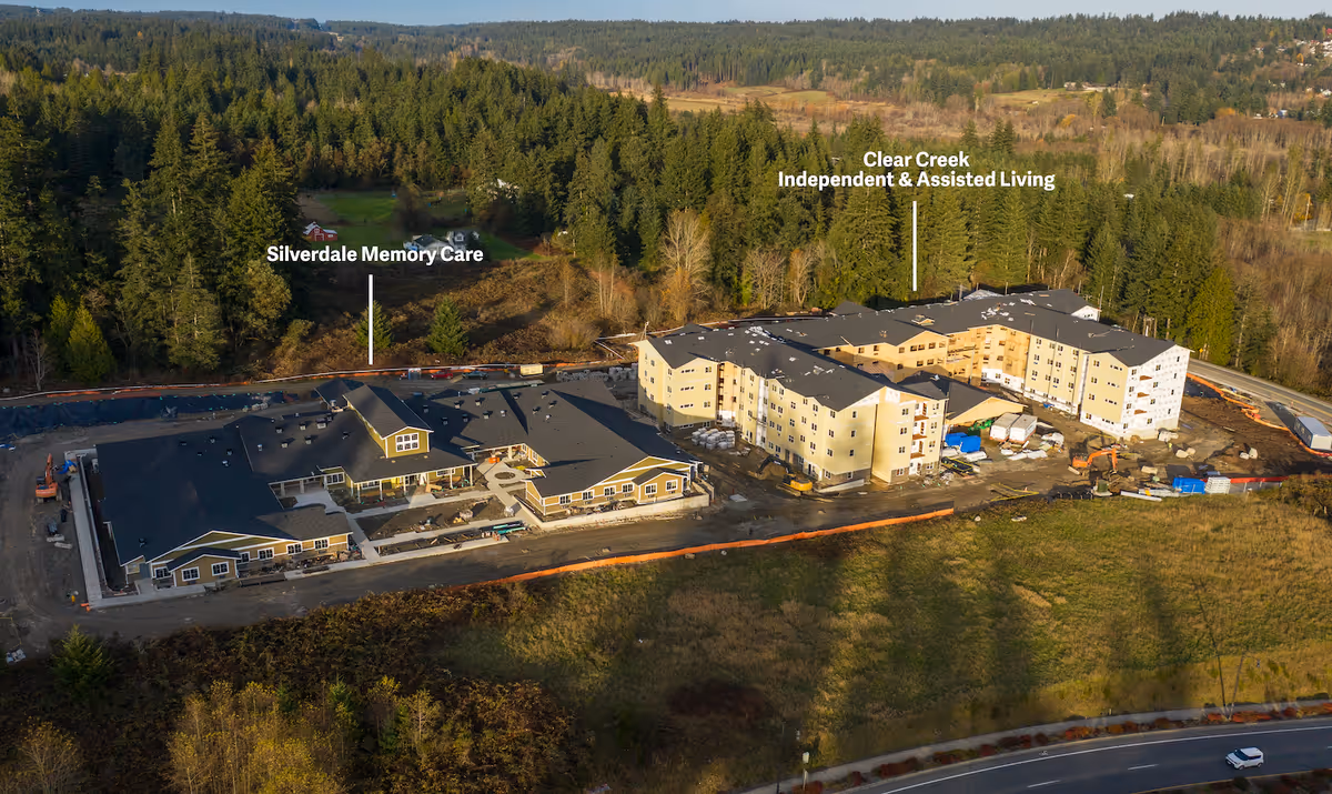 Aerial view of a large memory care and assisted living complex under construction surrounded by trees and fields, with labeled sections for Silverdale Memory Care and Clear Creek Independent & Assisted Living.