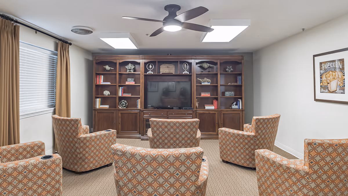 A cozy living room area with six patterned armchairs arranged in two rows facing a large wooden entertainment center with shelves holding books and decorative items. A flat-screen TV is centered on the entertainment unit. The room has beige carpet, a ceiling fan with light, two rectangular ceiling lights, a window with closed blinds and brown curtains on the left wall, and a framed popcorn-themed picture on the right wall.