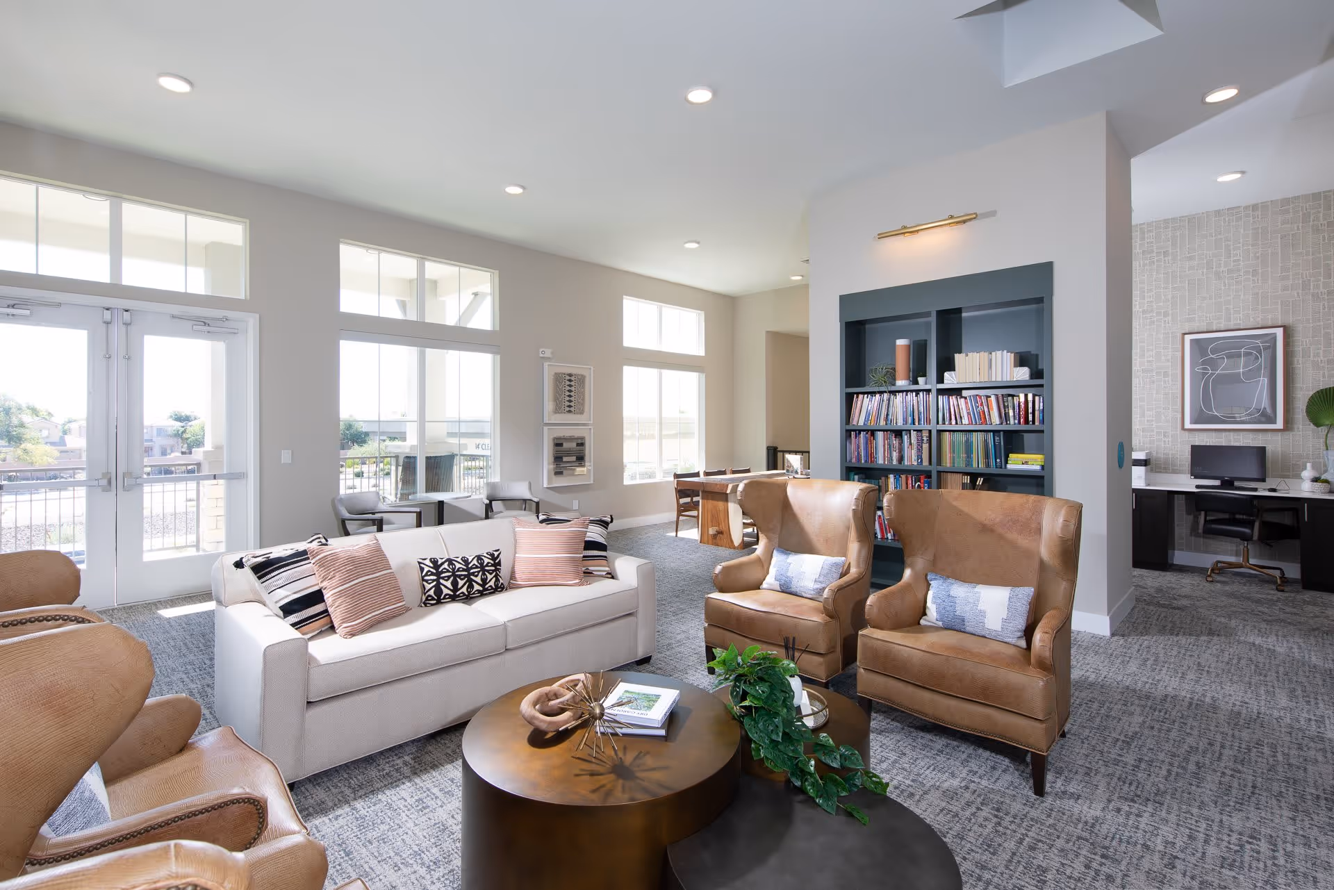 A bright and spacious living room area in a senior living facility with large windows letting in natural light. The room features a white sofa with patterned cushions, two brown leather armchairs with pillows, a round coffee table with decorative items and a plant, a bookshelf filled with books, and a desk with a computer in the background. The walls are painted light gray and the carpet is a textured gray pattern.