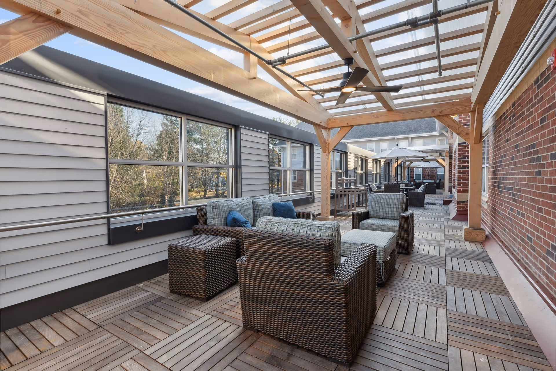 Outdoor patio area with wooden deck flooring and a wooden pergola overhead. The space is furnished with wicker chairs, cushioned sofas, ottomans, and side tables. Large windows line one side, and a brick wall lines the other. Ceiling fans are mounted under the pergola.