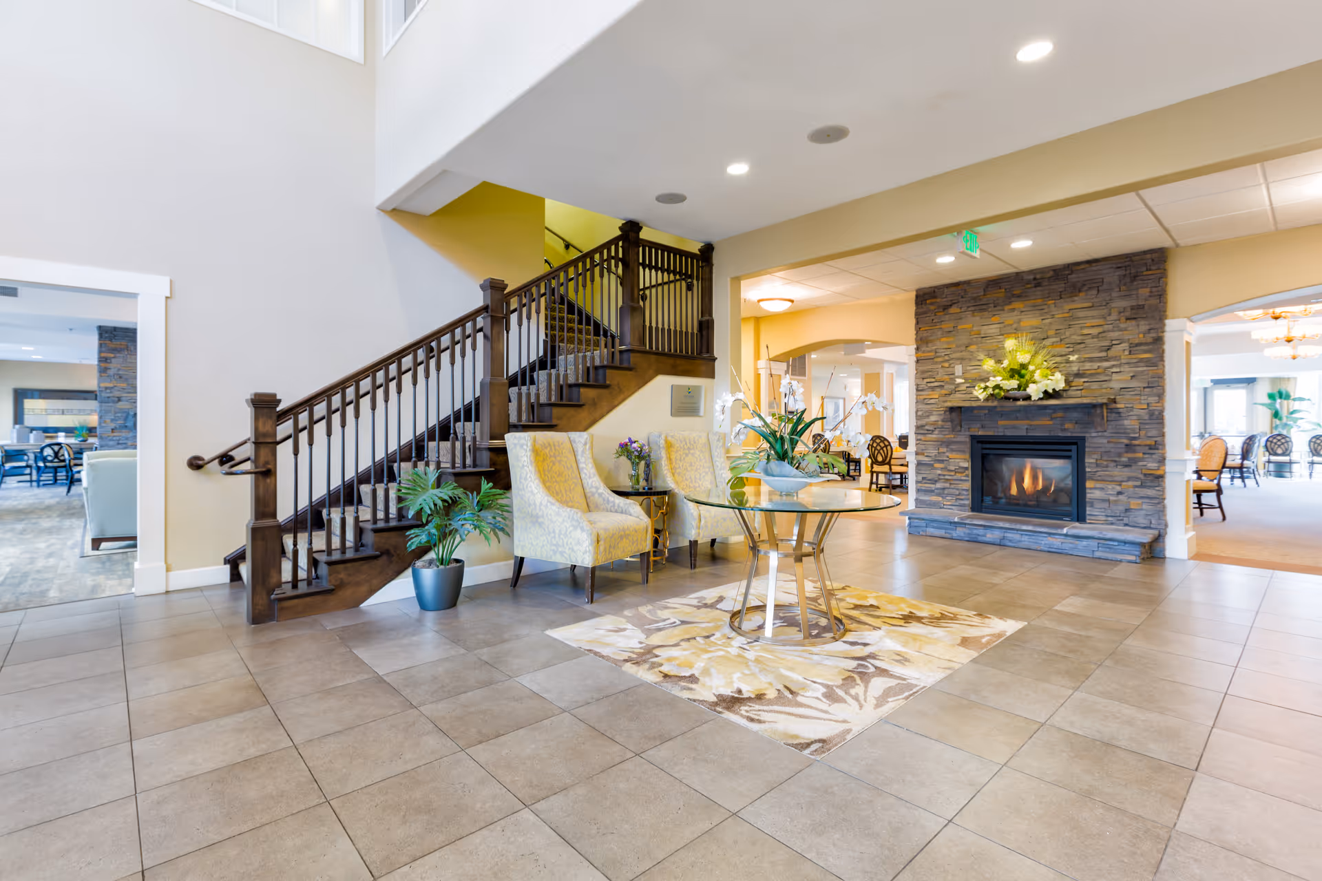 Spacious and well-lit interior of an assisted living facility featuring a tiled floor, a wooden staircase with dark railing, two upholstered chairs, a glass table with a floral centerpiece on a patterned rug, and a stone fireplace with a floral arrangement on the mantel. Adjacent rooms with additional seating and dining areas are visible.