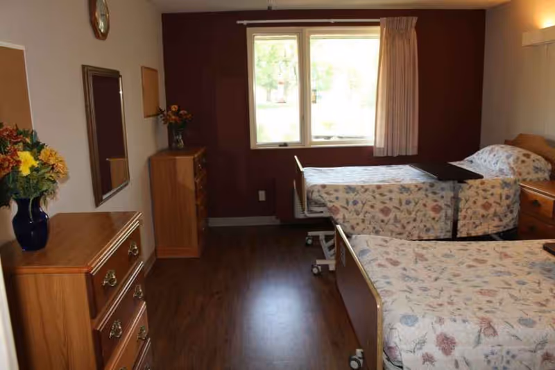 Two twin hospital-style beds with floral bedspreads in a senior facility bedroom with wooden dressers and a window.