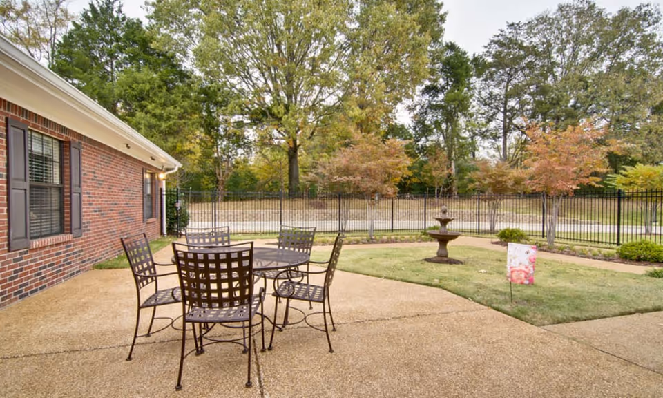 Patio area with a metal table and chairs on a paved courtyard beside a brick building, a small lawn with a fountain and trees beyond a fence.