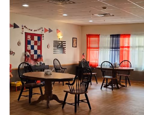 A cozy dining area with round wooden tables and black wooden chairs. The room is decorated with patriotic-themed items including a quilt on the wall, small flags, and red, white, and blue curtains covering the windows. The floor is wooden, and the lighting is warm with ceiling lights and a wall sconce.