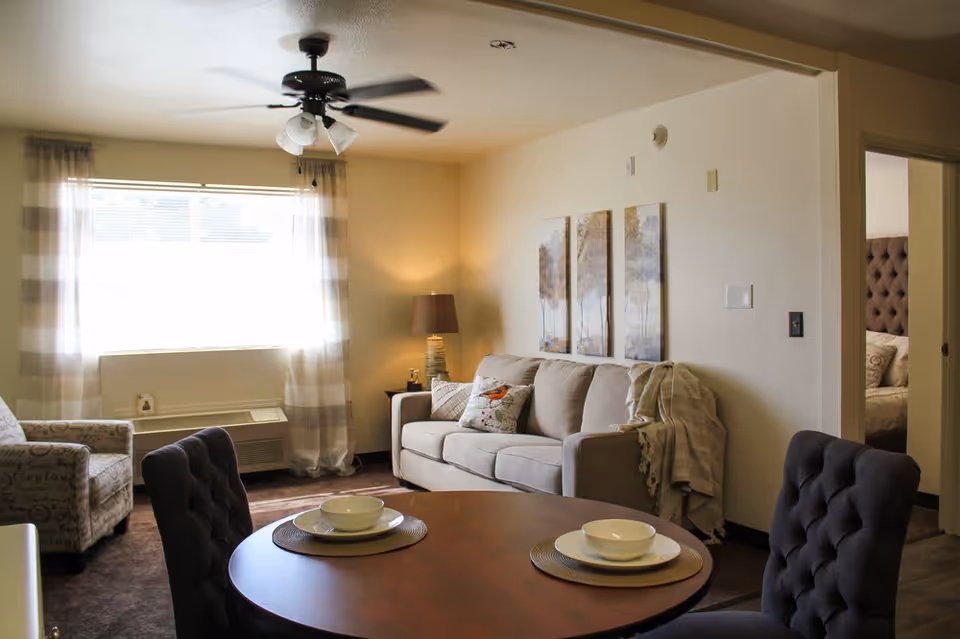 A cozy living room in a senior living facility with a beige sofa adorned with decorative pillows and a throw blanket, a patterned armchair, a round wooden dining table set with bowls and plates, a ceiling fan with lights, and a window with sheer striped curtains letting in natural light. A bedroom with a tufted headboard is visible through an open doorway.