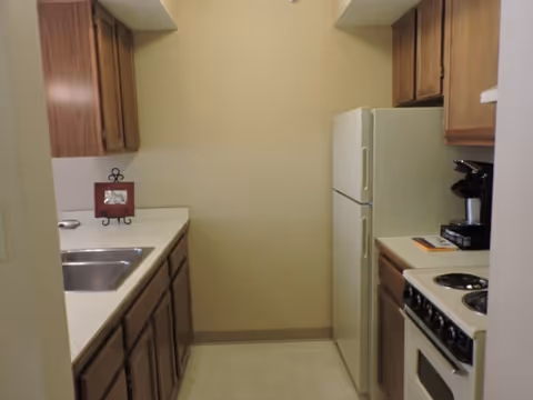 Small kitchen with wooden cabinets, a white refrigerator, a white stove with four burners, a stainless steel sink, and a coffee maker on the countertop.