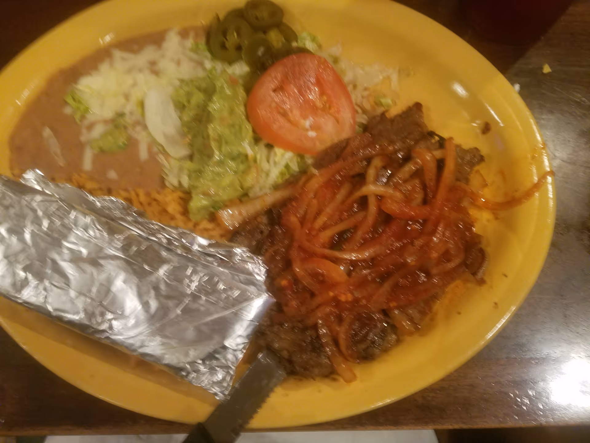Yellow plate with foil-wrapped burrito, salad, and saucy beef with onions on a wooden table.