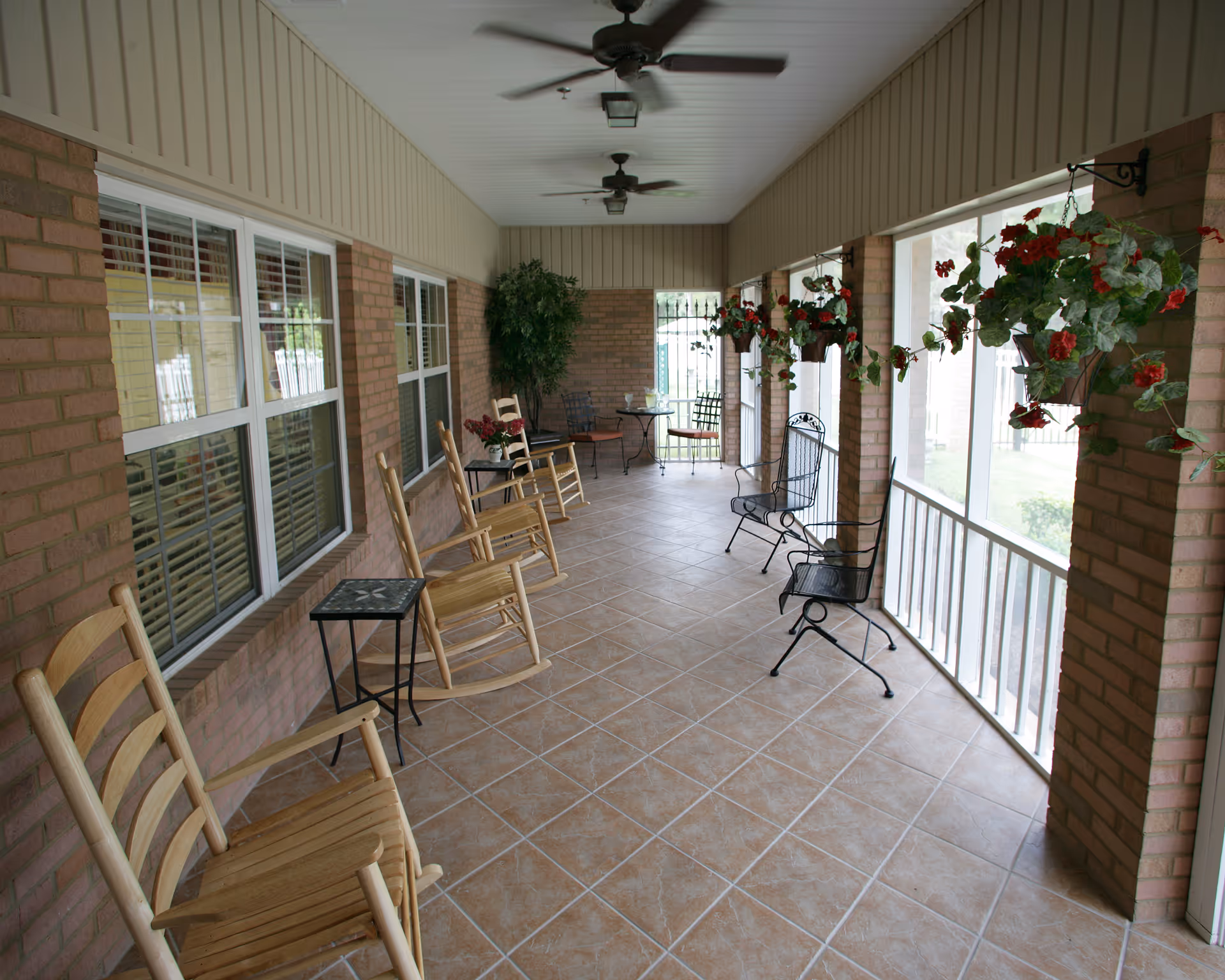 A covered outdoor patio area with tiled floor, wooden rocking chairs, metal chairs, small tables, hanging flower pots with red flowers, and ceiling fans. The patio is enclosed with brick pillars and screened windows.