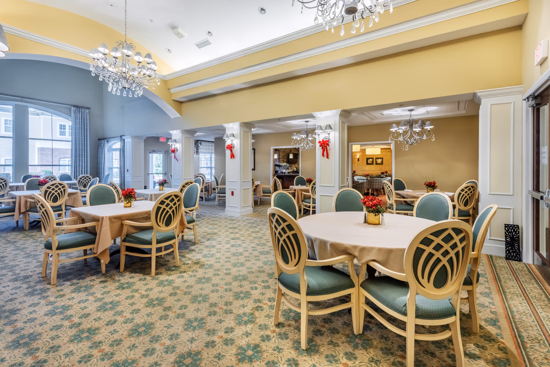 A spacious dining room with multiple round and rectangular tables covered with beige tablecloths, surrounded by chairs with green upholstery and decorative backs. The room features chandeliers hanging from a high ceiling with yellow and blue walls. There are red floral centerpieces on each table and red bows decorating the white columns. Large windows allow natural light to enter the room.