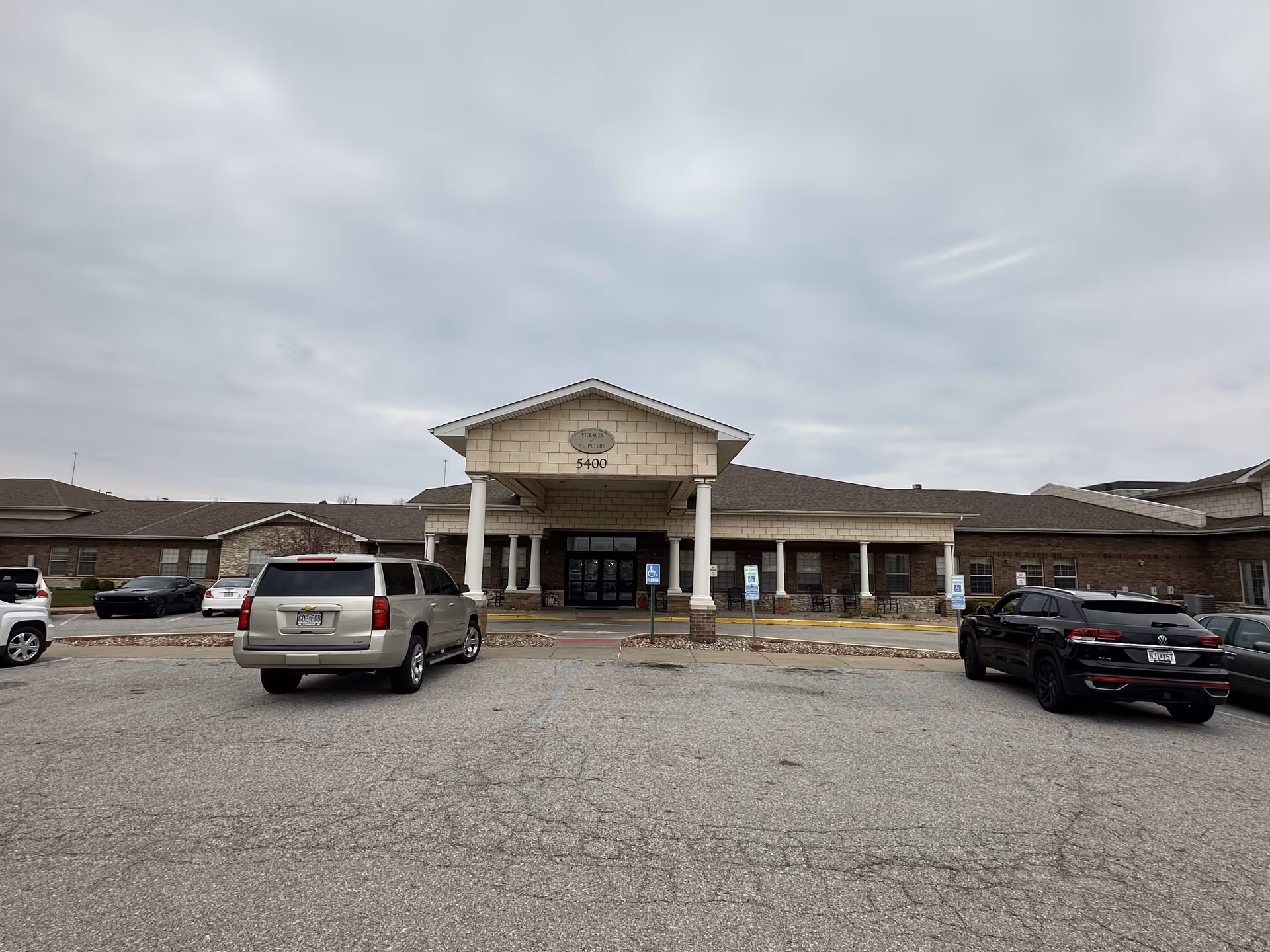 Front exterior view of the Villages of St. Peters facility showing a single-story building with a covered entrance supported by white columns. There are several parked cars in the parking lot in front of the building under a cloudy sky.