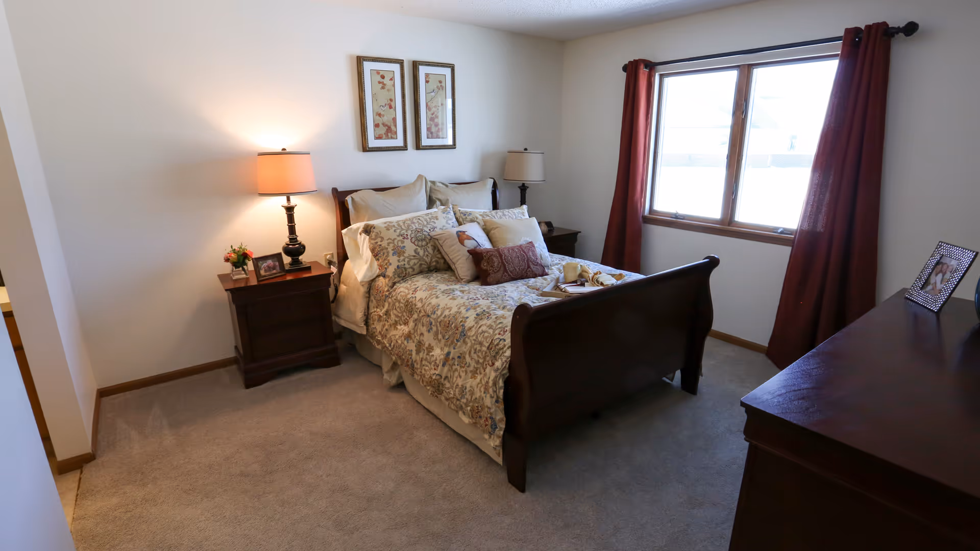 Bright furnished bedroom with a wooden sleigh bed, nightstands, a lamp and a window with red curtains.