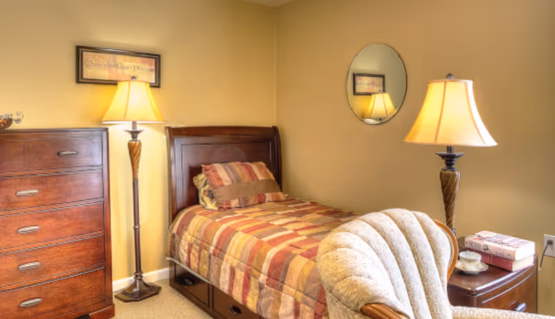 A cozy senior living bedroom with a single bed featuring a striped comforter and pillows. There is a wooden dresser with four drawers on the left, a floor lamp beside it, and a framed picture on the wall above. On the right side of the bed, there is a nightstand with a table lamp, a cup and saucer, and some books. A cushioned armchair is partially visible in the foreground. The walls are painted a warm beige color, and a round mirror hangs above the nightstand.