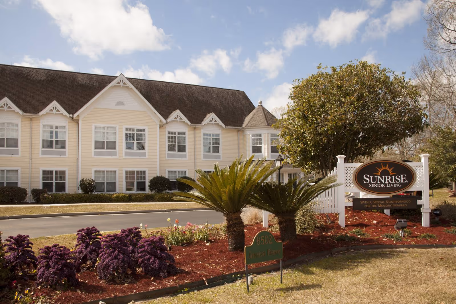 Exterior view of Sunrise Senior Living facility in Baton Rouge, showing a two-story building with multiple windows, a landscaped garden with purple flowers and palm plants, and a sign that reads 'Sunrise Senior Living' with a note about a special neighborhood for the memory impaired.