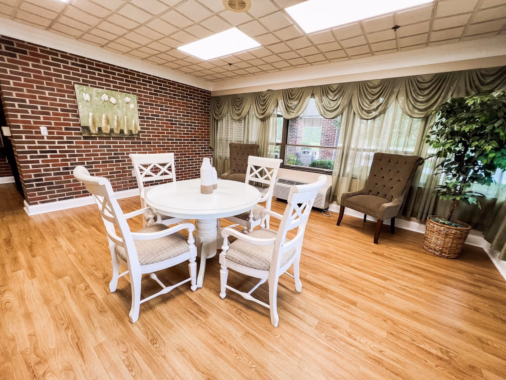 A bright room with a round white table surrounded by four white wooden chairs with cushioned seats. The room has a wooden floor, a brick accent wall with a painting of vases, large windows with green drapes, two upholstered armchairs, and a potted plant in a wicker basket.