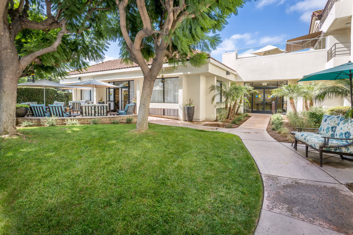 Courtyard with a green lawn, trees, patio seating and umbrellas leading to the assisted living building entrance.
